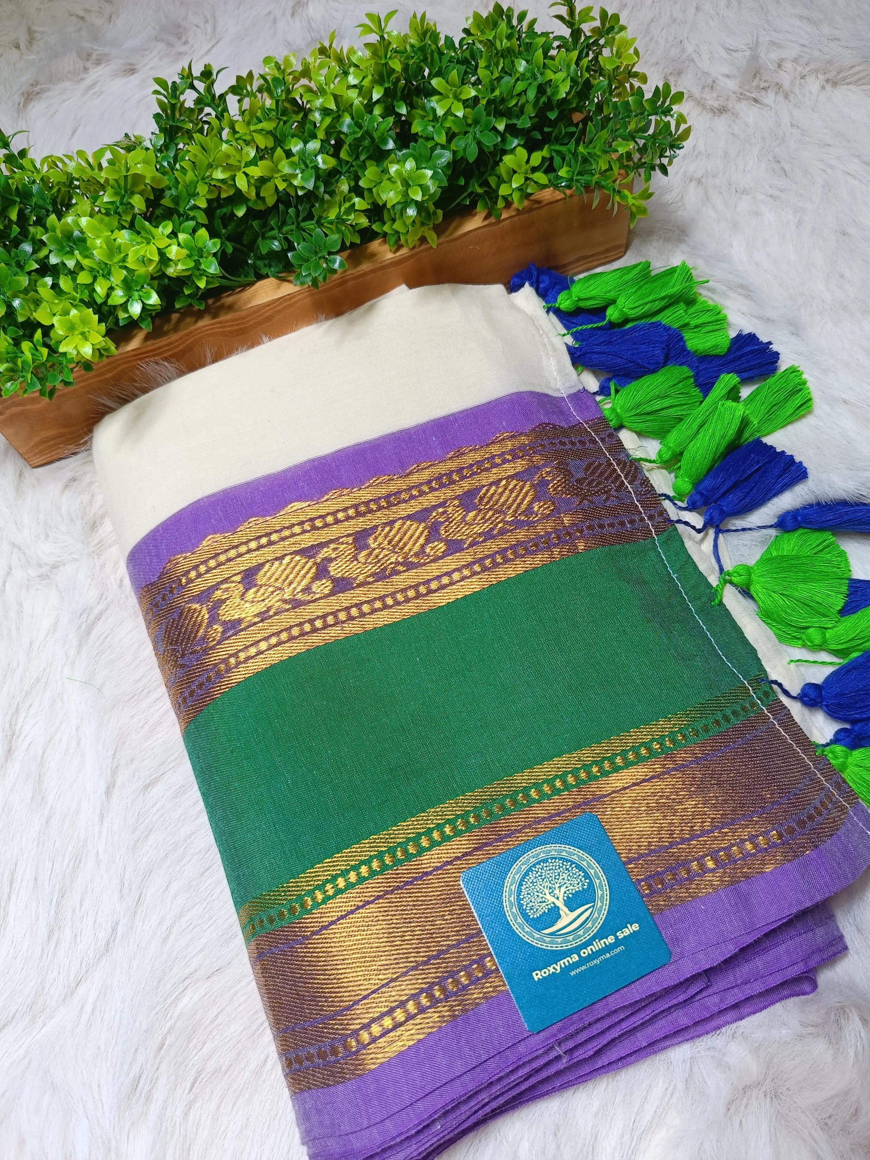 Green and purple fabric with gold patterns and a brand label, placed on a white surface with green plants in the background.